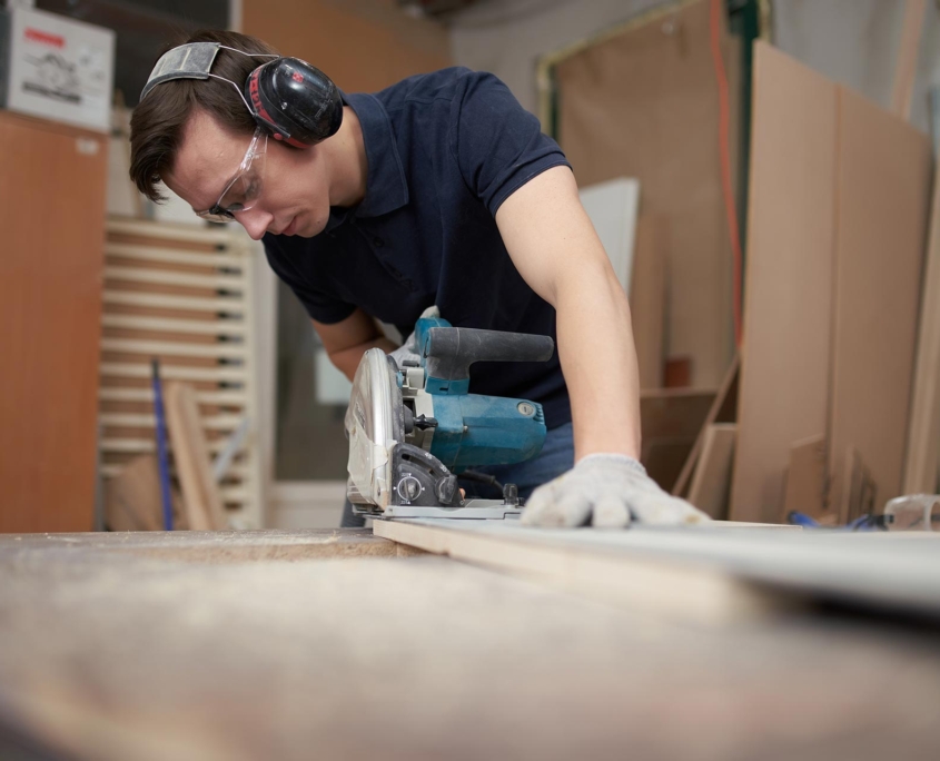 Carpenter man in protective headphones and white gloves works on jigsaw in workshop